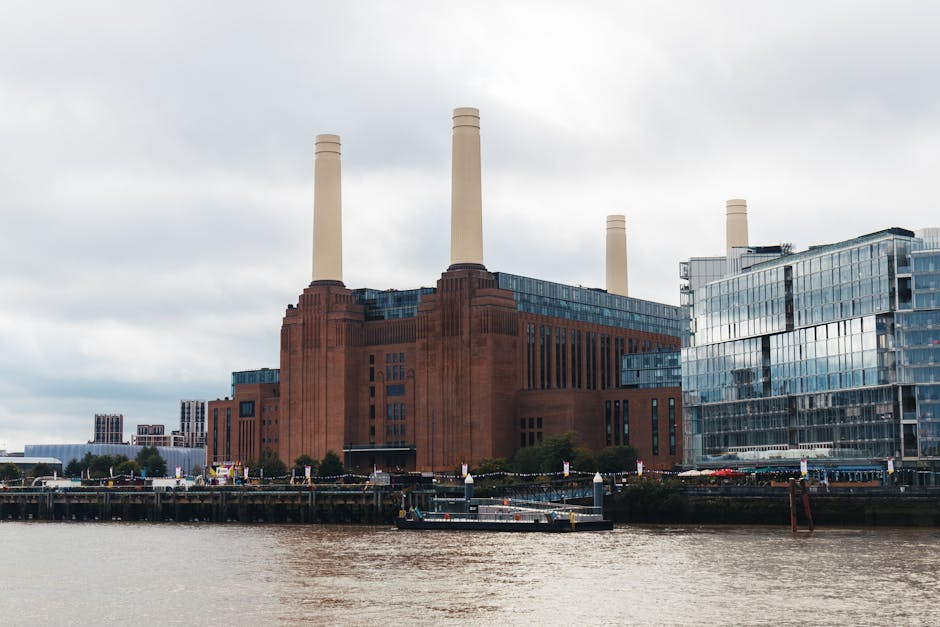 A large industrial building with multiple tall white chimneys, made of red brick and glass, situated along a waterway under a colorful, pink and purple sunset sky. The scene includes reflections of th